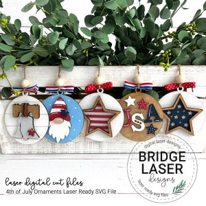 May include: A collection of five laser-cut wooden 4th of July ornaments. Each ornament features a unique design, including a Liberty Bell, a gnome, a star, and the letters "USA". They are decorated with red, white, and blue accents and hung with twine.