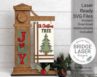 May include: A wooden sign display featuring a vertical post with a hanging rectangular panel. The post has a green inset with the red letters J and Y flanking a green wreath. The hanging panel displays the text Oh Christmas TREE above a green pine tree graphic. A small holly leaf accent with red berries sits at the base.