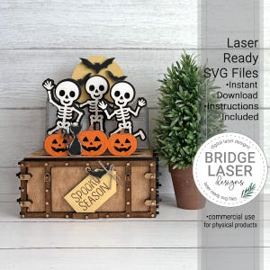 May include: A wooden box with a decorative topper featuring three white skeletons, three orange pumpkins, a black cat, and black bats against a yellow moon. A light-colored tag attached to the front of the box displays the text SPOOKY SEASON.