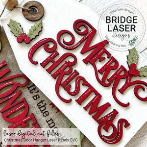 May include: A white wooden door hanger sign featuring red laser-cut lettering that reads Merry Christmas. The sign is decorated with green holly leaves and red berries. A circular wooden bead and a dark brown wooden ring are attached at the top.