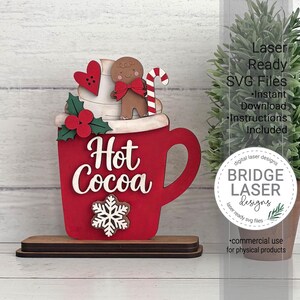 K&ouml;nnte beinhalten: Ein roter Holzausschnitt in Form einer Tasse mit dem wei&szlig;en Schriftzug Hot Cocoa, verziert mit einer Lebkuchenfigur, einem roten Herz, einer Zuckerstange und Stechpalmenbl&auml;ttern. Eine wei&szlig;e Schneeflocke ist an der Vorderseite der Tasse befestigt.