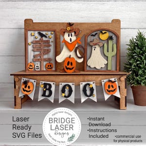 May include: A wooden tiered tray display set featuring laser-cut Halloween decorations. Includes a signpost with text, two ghost figures in cowboy hats, a cactus, and a banner spelling BOO with pumpkin accents. The set is crafted from wood.