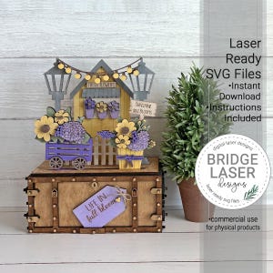 May include: A wooden laser-cut display featuring a yellow flower shop with purple and yellow flowers, a cart, and string lights. The base is a brown box with a purple tag reading "LIFE IN full bloom". Text includes "FRESH FLOWERS" and "SUNSHINE and BLOOMS".