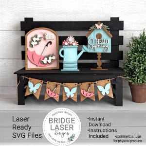 Puede incluir: Un banco de madera negro exhibe decoraciones de primavera cortadas con láser. Incluyen un paraguas rosa, una regadera azul y una casa para pájaros con el texto "Hello Spring". Una pancarta con mariposas y paraguas cuelga debajo. La imagen también incluye el texto "Laser Ready SVG Files".