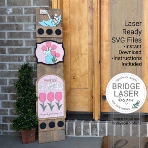 Puede incluir: Un letrero de madera con un diseño floral rosa y verde. El letrero dice "Farm Fresh Flowers" y "Pick your own". El letrero está decorado con una sombrilla azul, una maceta rosa y tres círculos negros. El letrero está apoyado contra una pared de ladrillo.