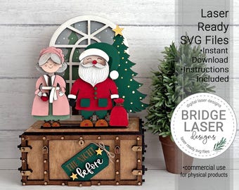 May include: A wooden laser-cut display featuring Santa Claus and Mrs. Claus standing before a window and a Christmas tree. The base is a rectangular box with a green tag labeled "THE NIGHT before" and two small yellow stars attached to the front.