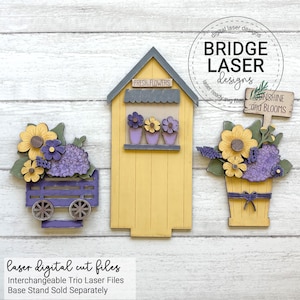 May include: Wooden laser-cut trio of flower-themed decorations. Includes a yellow flower shop with "FRESH FLOWERS" sign, a purple wagon with flowers, and a flower pot with a "Sunshine and Blooms" sign. The design features yellow, purple, and green flowers.
