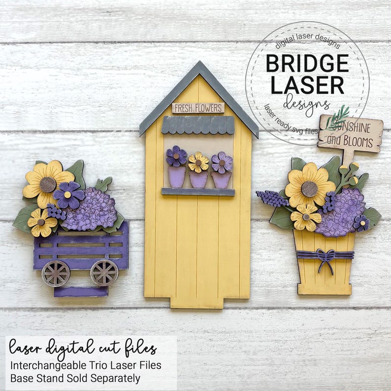 May include: Wooden laser-cut trio of flower-themed decorations. Includes a yellow flower shop with "FRESH FLOWERS" sign, a purple wagon with flowers, and a flower pot with a "Sunshine and Blooms" sign. The design features yellow, purple, and green flowers.