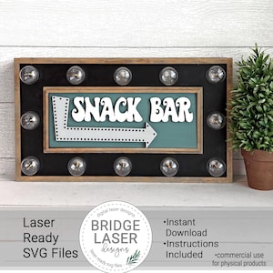 May include: A wooden sign with a black background and a teal blue sign inside. The sign has a white arrow pointing to the right and the words "SNACK BAR" in white letters. The sign has eight light bulbs around the perimeter.