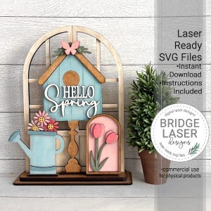 May include: A wooden spring-themed decoration featuring a birdhouse with "Hello Spring" text, a watering can, and a framed tulip design. The decoration is painted in pastel colours and includes a butterfly accent. The image also includes the text "Laser Ready SVG Files".
