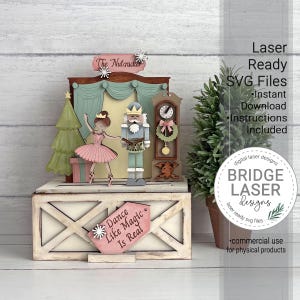 K&ouml;nnte beinhalten: Ein mehrschichtiges Holzdisplay mit einer Ballerina und einem Nussknacker auf einer B&uuml;hne, erg&auml;nzt durch einen Weihnachtsbaum und eine Standuhr. Die Basis tr&auml;gt das Schild "Dance Like Magic Is Real" und oben steht "The Nutcracker".