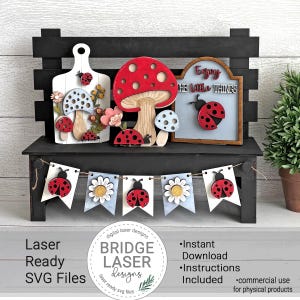 Op de afbeelding: Een zwart houten plank met lasergesneden houten decoraties van lieveheersbeestjes, paddenstoelen en bloemen. Een banner met lieveheersbeestjes en madeliefjes hangt eronder. Het bord luidt "Enjoy the little things". De afbeelding bevat ook de tekst "Laser Ready SVG Files".