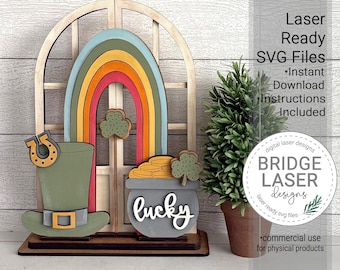 May include: A wooden St. Patrick's Day decoration featuring a rainbow, pot of gold, leprechaun hat, and horseshoe. The word "lucky" is written on the pot of gold. The text "Laser Ready SVG Files" is also visible.