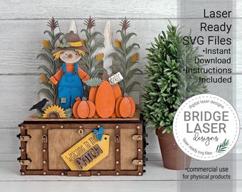 May include: A wooden laser-cut display featuring a scarecrow in blue overalls, orange pumpkins, and corn stalks standing on a rectangular box. A yellow tag attached to the front of the box reads Welcome to the PATCH with a small blue heart icon.