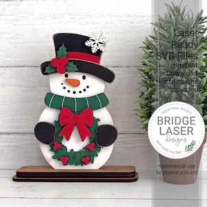May include: A layered wooden snowman decoration featuring a black top hat with a white snowflake, a green scarf, and a wreath with a red bow. The snowman has an orange carrot nose and black mittens. Text reads: Laser Ready SVG Files.