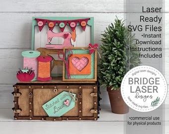 May include: A laser-cut wooden craft display featuring a sewing theme with a pink sewing machine, spools of thread, scissors, and a heart-shaped frame. A tag on the front reads Stitched with Love. Text includes Laser Ready SVG Files.
