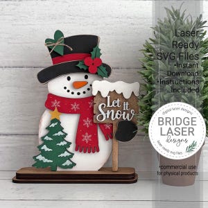 K&ouml;nnte beinhalten: Eine mehrschichtige Schneemann-Dekoration aus Holz mit schwarzem Zylinder, gr&uuml;nem Herz, roten Beeren, rotem Schal mit wei&szlig;en Schneeflocken, orangefarbener Nase und einem Schild mit der Aufschrift Let it Snow auf einem dunklen Sockel.
