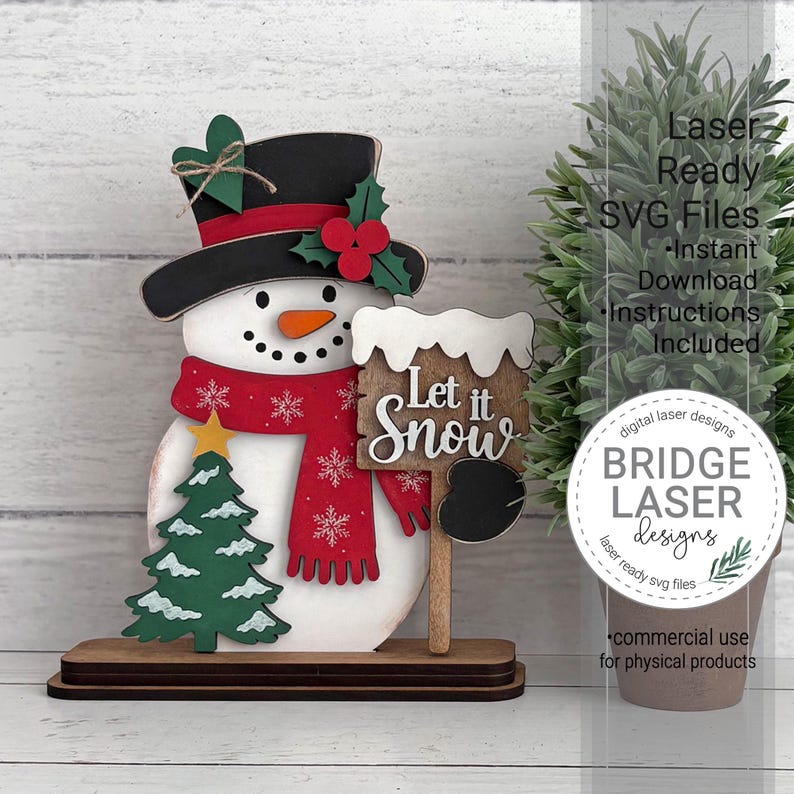 K&ouml;nnte beinhalten: Eine mehrschichtige Schneemann-Dekoration aus Holz mit schwarzem Zylinder, gr&uuml;nem Herz, roten Beeren, rotem Schal mit wei&szlig;en Schneeflocken, orangefarbener Nase und einem Schild mit der Aufschrift Let it Snow auf einem dunklen Sockel.