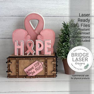 May include: A wooden tiered tray decor set featuring a pink awareness ribbon, two pink boxing gloves, and the word HOPE with a ribbon icon. A small pink tag attached to the base reads Never Stop the Fight. The set includes laser ready SVG files.