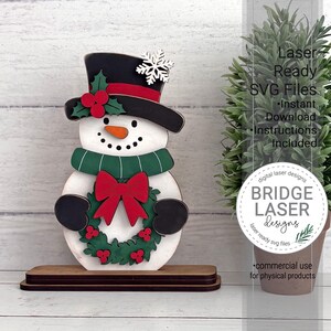 K&ouml;nnte beinhalten: Ein Schneemann-Ausschnitt aus Holz auf einem rechteckigen Sockel. Der Schneemann tr&auml;gt einen schwarzen Zylinder mit rotem Band, einer wei&szlig;en Schneeflocke und Stechpalmenbeeren. Er hat eine orangefarbene Karottennase, einen gr&uuml;nen Schal und einen Kranz.
