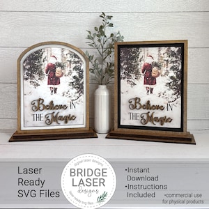 May include: Two wooden Christmas signs with the text "Believe in the Magic". One sign is arched, the other is rectangular. Both feature a Santa Claus image in a snowy forest. The signs are on a wooden base.