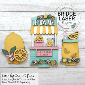 May include: Wooden laser-cut trio featuring a lemonade stand, a lemon, and a mason jar with a lemon slice. The stand is light blue with a yellow and white striped awning. The text "LEMONADE" and "FARM FRESH Lemons" are visible.