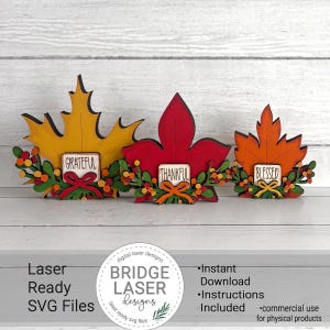 May include: Three wooden leaf-shaped decorations in yellow, red, and orange. Each leaf features a small rectangular sign with the words Grateful, Thankful, or Blessed, accented by green foliage, small colorful berries, and a small bow at the base.