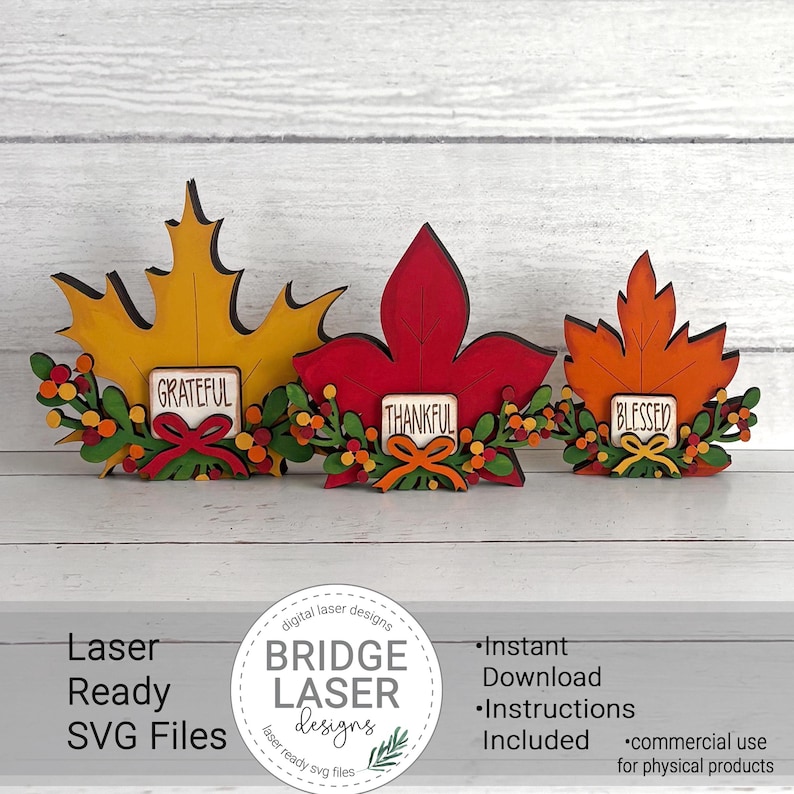 May include: Three wooden leaf-shaped decorations in yellow, red, and orange. Each leaf features a small rectangular sign with the words Grateful, Thankful, or Blessed, accented by green foliage, small colorful berries, and a small bow at the base.