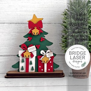 May include: A layered wooden Christmas tree decoration featuring a yellow star, red bows, and gold bells. Two gift boxes are at the base, one white with green trees and one red with white dots. Tags labeled Merry and Joy are attached to the bells.