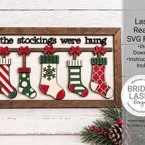 May include: A rectangular wooden sign with a dark brown frame and white background featuring the text 'the stockings were hung' in black. Five colorful Christmas stockings with red bows hang from a horizontal bar, each with unique patterns.