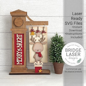 Op de afbeelding: Houten kerstdecoratie met een rendier met een rode sjaal, een cadeau en de woorden "MERRY & BRIGHT". Het bord bevat een bel, ster en ornament die boven het rendier hangen. Laserklare SVG-bestanden.