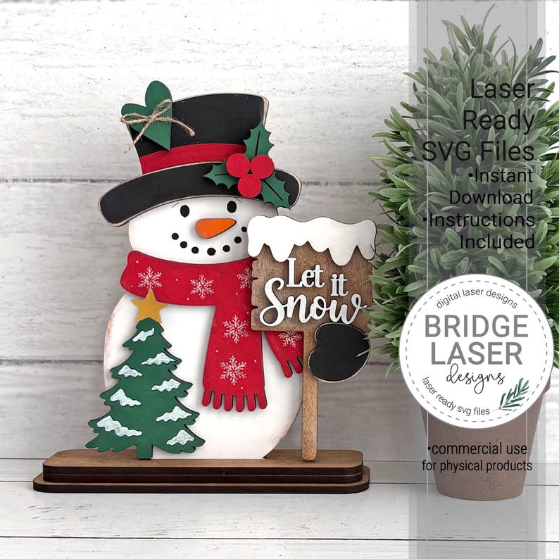 May include: A layered wooden snowman decoration featuring a black top hat with a green heart and red holly berries, a red scarf with white snowflakes, a green Christmas tree, and a sign with the text Let it Snow on a brown base.