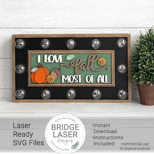 Op de afbeelding: Houten herfstbord met de tekst "I LOVE fall MOST OF ALL". Het bord heeft een pompoen, eikel en zonnebloem. Het bord is omgeven door gloeilampen en heeft de tekst "Laser Ready SVG Files".