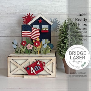 May include: A layered wooden craft display featuring a dark blue house with an American flag, red and blue fireworks, and colorful flowers. The base is a white farmhouse-style box with a red tag labeled 4th, accompanied by a small green potted tree.