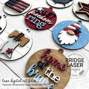 May include: A collection of patriotic wooden ornaments in red, white, and blue. Designs include an American flag, a gnome, and phrases like "Let Freedom Ring" and "Home of the Brave." Each ornament has a small hole for hanging.