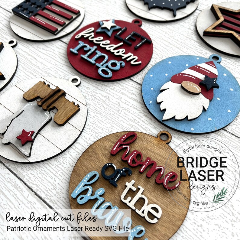 May include: A collection of patriotic wooden ornaments in red, white, and blue. Designs include an American flag, a gnome, and phrases like "Let Freedom Ring" and "Home of the Brave." Each ornament has a small hole for hanging.