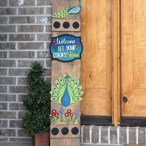Puede incluir: Un letrero de bienvenida de madera con un diseño de pavo real. El letrero presenta el texto "Welcome Let Your Colors Shine" y tiene un pavo real en la parte superior e inferior. El letrero está decorado con flores coloridas y se coloca junto a una pequeña planta en maceta.