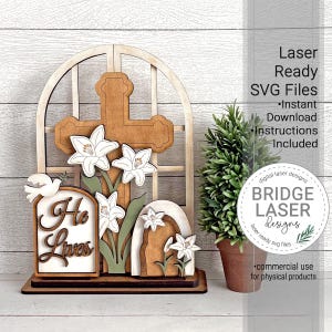 May include: A wooden Easter decoration featuring a cross, lilies, and the words "He Lives." The design includes a dove and an arched structure. The text "Laser Ready SVG Files" and "Bridge Laser Designs" are also visible.