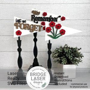 Könnte beinhalten: Drei weiße Holz-Wimpel mit schwarzer Beschriftung und roten Mohnblumen. Die Wimpel tragen die Aufschriften "We Remember", "Last We Forget" und eine Ansammlung roter Mohnblumen. Die Wimpel stehen auf schwarzen Holzständern.