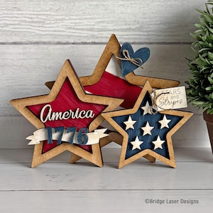 May include: Wooden star decorations in patriotic colours. One star features a red interior with the text "America 1776", another has a blue interior with white stars, and a third has a blue heart. The decorations are on a white wooden surface.