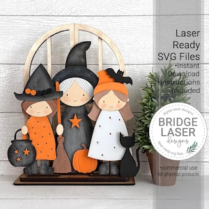 Puede incluir: Decoración de Halloween de madera con tres figuras de brujas, un gato negro y una calabaza. Las brujas visten vestidos y sombreros naranjas, negros y blancos. La decoración incluye el texto "Laser Ready SVG Files" y "Bridge Laser Designs".