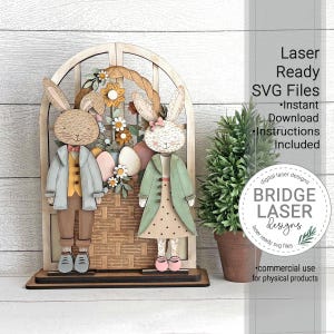 May include: Wooden Easter decoration with two bunny figures in coats, positioned before a basket of flowers and Easter eggs, set within a window frame. Text on the right reads "Laser Ready SVG Files".