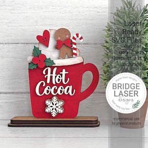 May include: A red wooden mug cutout with the text Hot Cocoa in white, topped with a gingerbread person, a red heart, a candy cane, and holly berries. A white snowflake is attached to the front, and the entire piece stands on a wooden base.