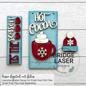 K&ouml;nnte beinhalten: Ein Set aus drei lasergeschnittenen Holzschildern mit Winterthema. Die Schilder zeigen den Text "Tis the season", "Hot Cocoa" und ein Schneeflocke. Die Schilder sind in Rot, Wei&szlig; und Blau bemalt.