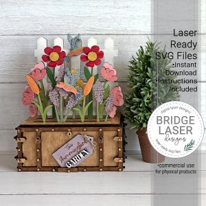 K&ouml;nnte beinhalten: Eine Holzbox mit einer lasergeschnittenen Gartenszene, bestehend aus einem wei&szlig;en Lattenzaun, roten und rosa Blumen sowie zwei Kolibris. Ein lila Schild an der Vorderseite tr&auml;gt die Aufschrift The Hummingbird GARDEN. Text rechts: Laser Ready SVG Files.