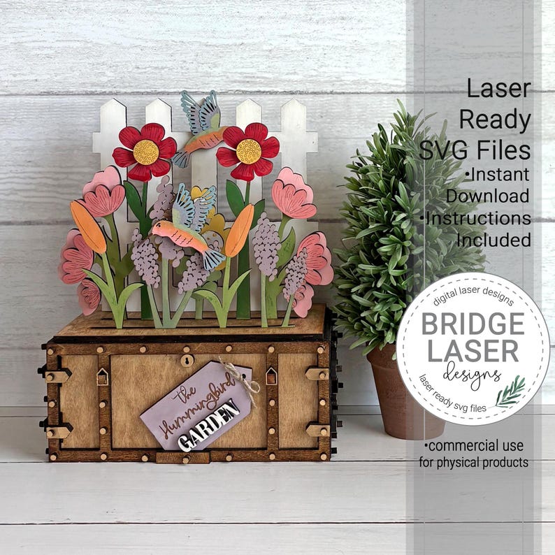 K&ouml;nnte beinhalten: Eine Holzbox mit einer lasergeschnittenen Gartenszene, bestehend aus einem wei&szlig;en Lattenzaun, roten und rosa Blumen sowie zwei Kolibris. Ein lila Schild an der Vorderseite tr&auml;gt die Aufschrift The Hummingbird GARDEN. Text rechts: Laser Ready SVG Files.