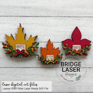 May include: Three decorative wooden leaf cutouts in yellow, orange, and red. Each leaf features a small rectangular sign with the words Grateful, Blessed, or Thankful, accented by green foliage and small colorful berries at the base of each leaf.