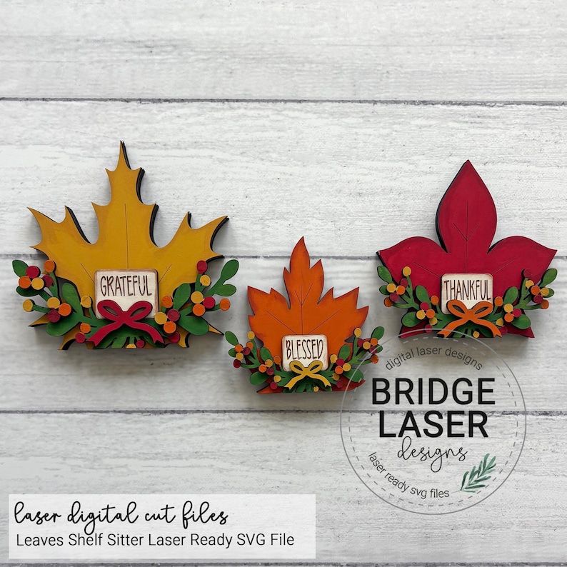 May include: Three decorative wooden leaf cutouts in yellow, orange, and red. Each leaf features a small rectangular sign with the words Grateful, Blessed, or Thankful, accented by green foliage and small colorful berries at the base of each leaf.