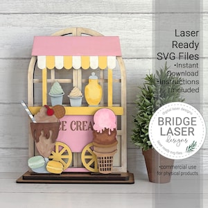 May include: A wooden ice cream cart with a pink awning and yellow accents. The cart features ice cream cones, sundaes, and macarons. The text "ICE CREAM" is visible on the cart. The image also includes the text "Laser Ready SVG Files" and "Bridge Laser Designs".
