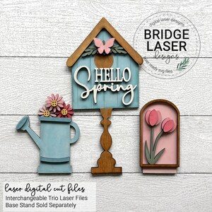 Puede incluir: Un tr&iacute;o de decoraciones de madera cortadas con l&aacute;ser con tem&aacute;tica primaveral. Incluye una casita para p&aacute;jaros azul con el texto "HELLO SPRING", una regadera con flores rosas y un dise&ntilde;o de tulipanes enmarcados. La casita tiene un acento de mariposa.